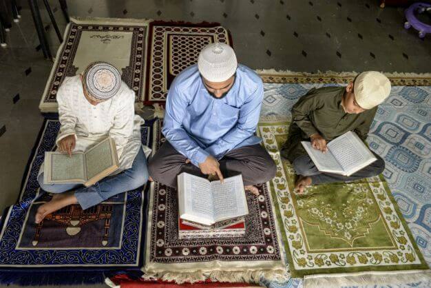A group of young students sitting in a circle in a brightly lit classroom, attentively listening to an Islamic scholar teaching Quranic recitation, with open Qurans and notebooks in front of them.
